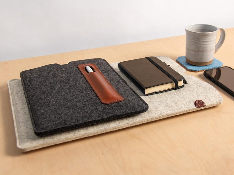 Puede incluir: Una funda de fieltro para port&aacute;til con un portal&aacute;pices de cuero, un cuaderno marr&oacute;n y una taza de cer&aacute;mica sobre una superficie de madera. Tambi&eacute;n hay un smartphone y un posavasos azul. La funda es de color gris oscuro y el cuaderno tiene un cierre el&aacute;stico negro.