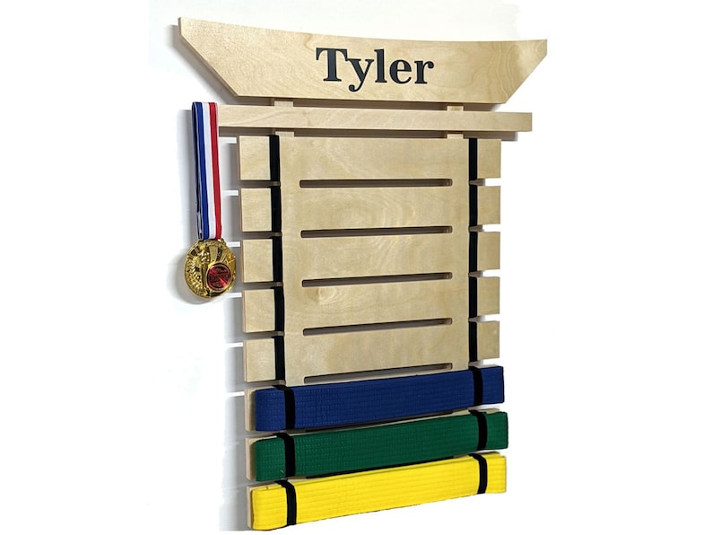 Pu&ograve; includere: Un porta cintura per arti marziali in legno con il nome "Tyler" in cima. Il supporto ha fessure per le cinture e un gancio per una medaglia. Ci sono quattro cinture sul supporto: blu, verde, giallo e nero.