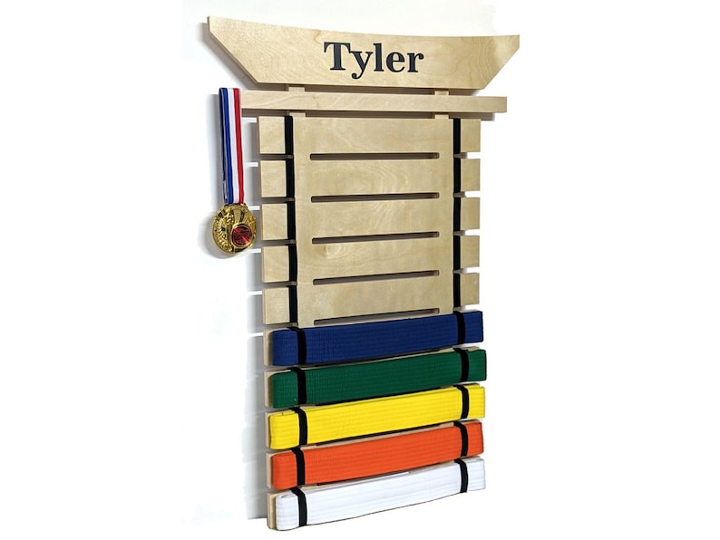 May include: A wooden martial arts belt display with the name "Tyler" at the top. The display holds several colored belts: white, orange, yellow, green, and blue. A gold medal hangs on the side.