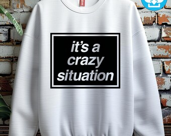 May include: White sweatshirt with the phrase "it's a crazy situation" in a black rectangle. The text is in a silver, sans-serif font. The sweatshirt has a crew neck and long sleeves. A small red tag is visible near the collar.