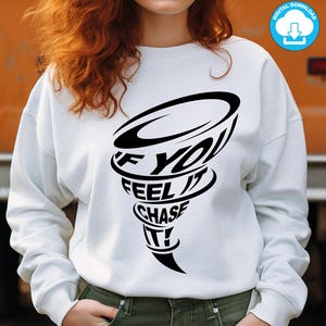 Puede incluir: Sudadera blanca con un gráfico negro de un tornado con el texto "If you feel it chase it!" impreso en ella.