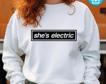 May include: White sweatshirt with the text "she's electric" in a black rectangle. The text is in a white, sans-serif font. The sweatshirt has long sleeves and a crew neck.