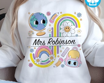 May include: White sweatshirt with a colorful school-themed design. Features a globe, rainbow, smiling flower, pencil, backpack, and the name "Mrs. Robinson" in black script. Includes a digital download icon.
