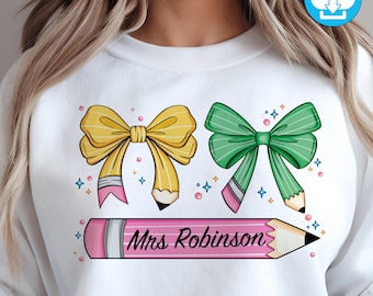 May include: White sweatshirt with a pink pencil design and two bows, one yellow and one green. The pencil has the text "Mrs. Robinson" written on it.