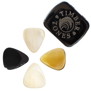 May include: A set of four guitar picks in various colors, including black, cream, and amber, arranged around a black case. The case is square with rounded corners and features the text "TIMBER TONES" and a decorative emblem.