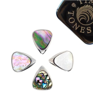 Può includere: Quattro plettri per chitarra con intarsi di conchiglia di abalone iridescente e bordi in metallo color argento. Un astuccio nero con testo dorato è nell'angolo in alto a destra. I plettri sono disposti a cerchio.