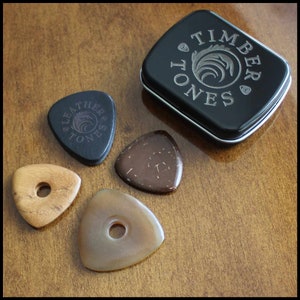 This image shows the 4 Picks we have chosen for Bass Guitar. A large black 3mm thick leather, a triangle coconut husk pick, a triangular Indian Teak pick with a hole through the middle and a large Clear Horn pick. They are next to the black tin.