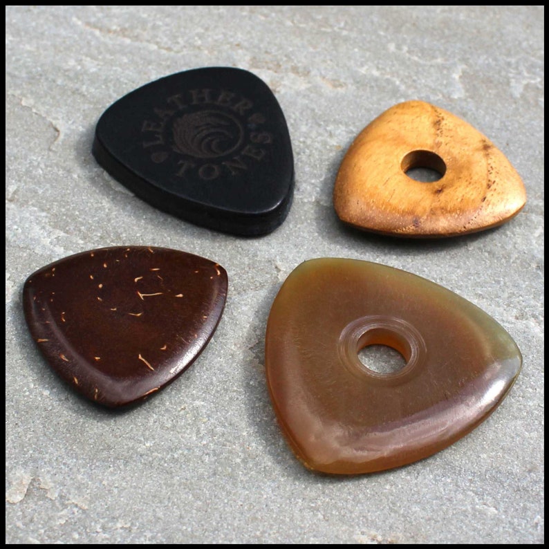 This image shows the 4 Picks we have chosen for Bass Guitar. A large black 3mm thick leather, a triangle coconut husk pick, a triangular Indian Teak pick with a hole through the middle and a large Clear Horn pick. They are on a stone background.