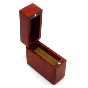 May include: Open, rectangular wooden box with a hinged lid. The box is made of reddish-brown wood and has a light-colored interior lining. A small, round, gold-colored clasp secures the lid.
