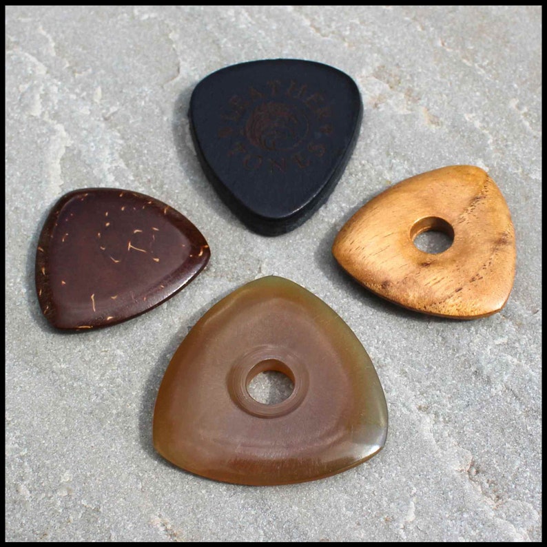 This image shows the 4 Picks we have chosen for Bass Guitar. A large black 3mm thick leather, a triangle coconut husk pick, a triangular Indian Teak pick with a hole through the middle and a large Clear Horn pick. They are on a stone background.