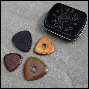 This image shows the 4 Picks we have chosen for Bass Guitar. A large black 3mm thick leather, a triangle coconut husk pick, a triangular Indian Teak pick with a hole through the middle and a large Clear Horn pick. They are on a stone background.