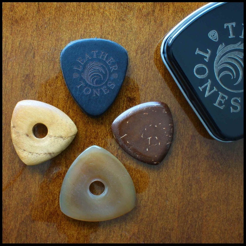 This image shows the 4 Picks we have chosen for Bass Guitar. A large black 3mm thick leather, a triangle coconut husk pick, a triangular Indian Teak pick with a hole through the middle and a large Clear Horn pick. They are next to the black tin.