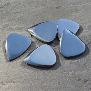 Op de afbeelding: Vijf glanzende, zilverkleurige gitaar plectrums op een grijze, getextureerde ondergrond. De plectrums hebben een glad, reflecterend oppervlak en een druppelvorm, met een licht afgeronde punt. De plectrums zijn van metaal.