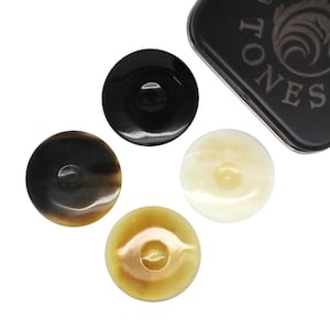May include: Four round guitar picks in varying colors: black, dark brown, cream, and light brown. The picks have a glossy finish and a hole in the center. A black metal container with the word "TONES" is partially visible.