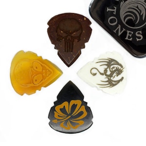 May include: Four guitar picks with different designs. The picks are made of different materials, including wood, plastic, and bone. The designs include a skull, a dragon, a flower, and an alien. The picks are arranged in a cross shape on a white background.
