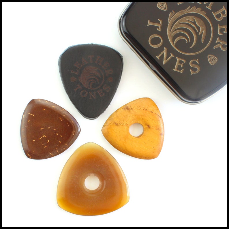 This image shows the 4 Picks we have chosen for Bass Guitar. A large black 3mm thick leather, a triangle coconut husk pick, a triangular Indian Teak pick with a hole through the middle and a large Clear Horn pick. They are on a white background.
