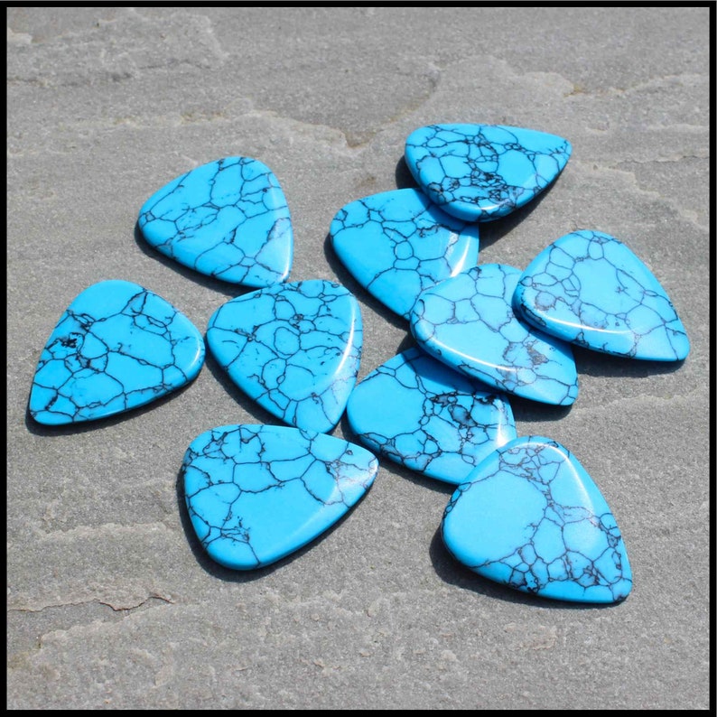 This image show 5 Blue Dragon Skin Reconstituted Stone Guitar Picks. These plectrums have black veins running through a blue background. They are randomly arranged on a stone back ground so you can see the 3D shape of their design.