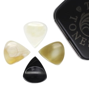 Puede incluir: Cuatro púas de guitarra en crema, blanco roto y ámbar están dispuestas alrededor de una caja de metal negro. Las púas tienen una forma suave y redondeada. La caja es negra con texto blanco y un logotipo.