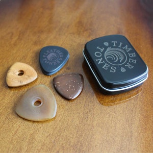May include: A collection of guitar picks in various colors and materials, including wood and stone. A black metal tin with the words "Timber Tones" is also present. The picks are arranged on a wooden surface.