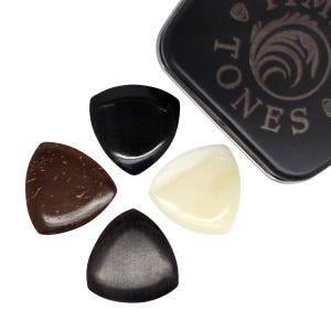 May include: Four guitar plectrums in different materials and colours, including black, brown, and cream, arranged around a black metal case. The case has the text "TONES" and a decorative design.