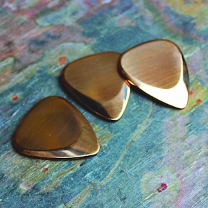 Può includere: Tre plettri per chitarra dorati, a forma di lacrima, sono disposti in fila su una superficie testurizzata blu e marrone.