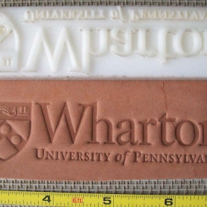 May include: A white rubber stamp with the text "University of Pennsylvania Wharton" and a crest. The stamp is on top of a brown leather surface with the same text and crest embossed into the leather.