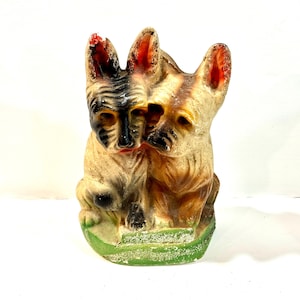 May include: A vintage-style molded figurine featuring two dogs sitting side-by-side on a green base. The dog on the left has black and tan markings, while the dog on the right is primarily tan with dark accents. Both have pointed ears and textured fur.