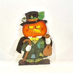Puede incluir: Decoración de Halloween de madera con una cabeza de calabaza con nariz de zanahoria tallada, que lleva un sombrero de copa negro y un traje verde. La figura sostiene un bastón y un saco marrón, con un pequeño ratón en el sombrero.