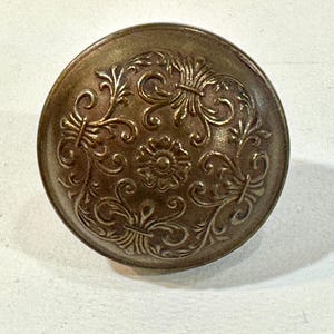 May include: A round, antique-style button with an ornate floral design. The button is a bronze color and features a raised pattern of swirling leaves and a central flower. The button is likely made of metal and is suitable for sewing or crafting projects.