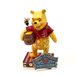Könnte beinhalten: Eine gelbe Bärenfigur in rotem Shirt hält einen braunen Honigtopf mit der Aufschrift HUNNY, über dem eine kleine Biene schwebt. Der Bär steht auf einem Sockel mit Blumenmuster und einem hängenden Disney Traditions Jim Shore Enesco Etikett.