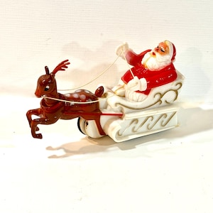 Könnte beinhalten: Eine Kunststofffigur im Vintage-Stil, die ein braunes Rentier mit weißen Flecken zeigt, das einen weißen Schlitten mit Goldverzierungen zieht. Der Weihnachtsmann mit rotem Anzug und weißem Bart sitzt in dem Schlitten.