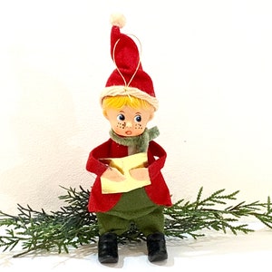 Puede incluir: Una pequeña figura de elfo con cabello rubio, pecas y ojos azules, que lleva un gorro rojo con un pompón blanco, una chaqueta roja, una bufanda verde y pantalones verdes. El elfo sostiene un libro dorado frente a una rama de pino verde.