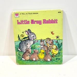 May include: A vintage Tell-A-Tale book titled Little Gray Rabbit with a light green cover. The illustration features a large gray rabbit gesturing toward a group of smaller rabbits and yellow chicks in a grassy field with flowers and a butterfly.
