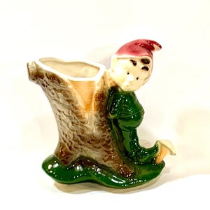 May include: A ceramic planter shaped like a tree stump with a smiling elf figure. The elf wears a green outfit and a red hat. The planter has a beige interior and a brown exterior with green accents.