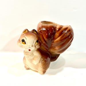 Può includere: Un vaso in ceramica a forma di scoiattolo con una finitura marrone lucida sulla coda e un colore marrone chiaro sul corpo. Lo scoiattolo ha occhi neri dipinti, una piccola bocca rossa e un'apertura cava sul retro per contenere piccoli oggetti.