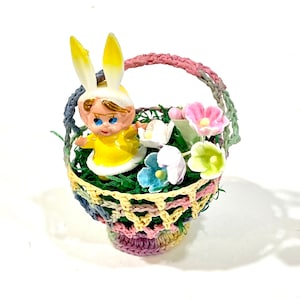 May include: A small, colorful Easter basket with a crocheted design. The basket contains a figurine of a child wearing bunny ears, surrounded by artificial flowers in pink, white, blue, and green. The basket handle is pink.