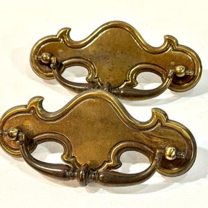 May include: Two antique brass drawer pulls with ornate, curved designs. Each pull features a decorative backplate and a curved handle. The brass has a warm, aged patina, suggesting vintage or antique furniture hardware.