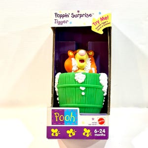 May include: A Poppin' Surprise Tigger toy by Mattel in original packaging. The orange and black striped Tigger character is emerging from a green barrel with white foam accents. The box features the text Poppin' Surprise Tigger and Pooh.
