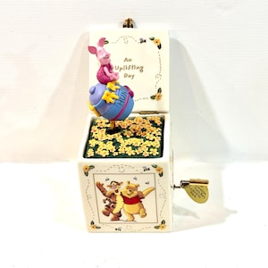 May include: A ceramic music box featuring a Piglet figurine sitting on a blue honey pot labeled HUNNY. The lid interior reads An Uplifting Day. The front panel displays a graphic of Tigger and Winnie the Pooh. A gold metal crank is on the side.