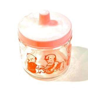 Pode incluir: Um frasco de vidro transparente com uma tampa de plástico rosa claro com um botão central. O exterior do recipiente de vidro é decorado com ilustrações gráficas cor de laranja de dois bebés sentados a brincar com pequenos objetos redondos.