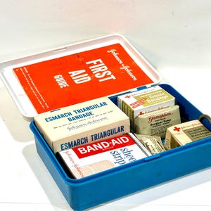 May include: A blue first aid kit with a white lid, containing bandages, gauze, and a first aid guide. The kit includes Band-Aid brand adhesive strips, triangular bandages, and surgical soap. The lid has the text "FIRST AID GUIDE".