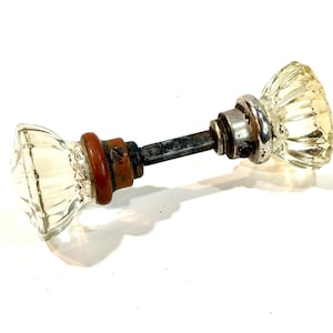 May include: A clear, faceted glass door knob set featuring two octagonal glass handles connected by a central metal spindle. One side includes a dark reddish-brown circular base, while the other side features a silver-toned metal collar with a small screw.