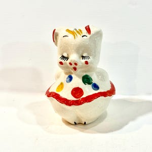 Op de afbeelding: Een vintage keramieken varkensbeeldje met gesloten ogen, rode accenten en een polkadot jurk. Het varken heeft gele en rode details op zijn hoofd en een rode rand op de jurk. Het beeldje is wit met een craquelé-afwerking.