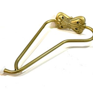 May include: A brass-toned metal wall hook featuring a decorative, ornate mounting plate with a fan-like center design. The hook has a triangular frame structure made of curved metal tubing and terminates in a small, smooth white spherical tip.