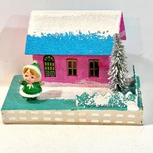 May include: A vintage Christmas diorama featuring a pink house with a blue roof and snow-covered trim. A small figurine in a green outfit stands in front of the house. A small Christmas tree and fence are also present.