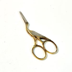 May include: Gold-toned sewing scissors with a crane design. The scissors feature a detailed crane body with etched silver-colored wings and a long, slender beak. The handles are oval-shaped and the blades are silver.