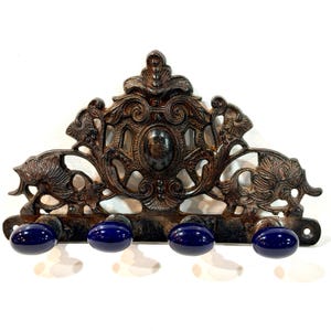 May include: A wall-mounted coat rack featuring a dark, weathered bronze-toned metal frame with intricate scrollwork and floral motifs. The rack includes four horizontal mounting holes and four protruding hooks, each tipped with a smooth, oval-shaped, deep cobalt blue ceramic knob.