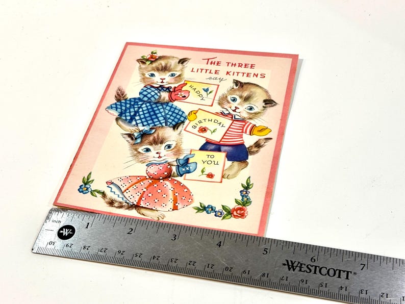 May include: Vintage birthday card featuring three kittens holding signs that read "Happy Birthday to You." The kittens are dressed in blue and red outfits. The card has a pink border and floral accents.