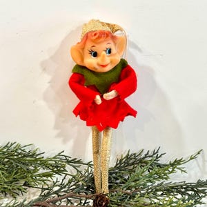 May include: A vintage Christmas elf ornament. The elf has a painted face with rosy cheeks, blue eyes, and a gold glitter hat. It wears a red dress with a green collar and gold glitter legs. The ornament is displayed on a bed of greenery.