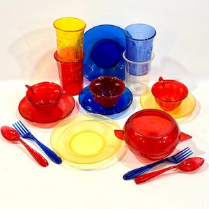 May include: A collection of translucent plastic toy dishware in primary colors including red, blue, and yellow. The set features cups, saucers, plates, a lidded bowl, spoons, and forks. The items are arranged in a group on a white surface.